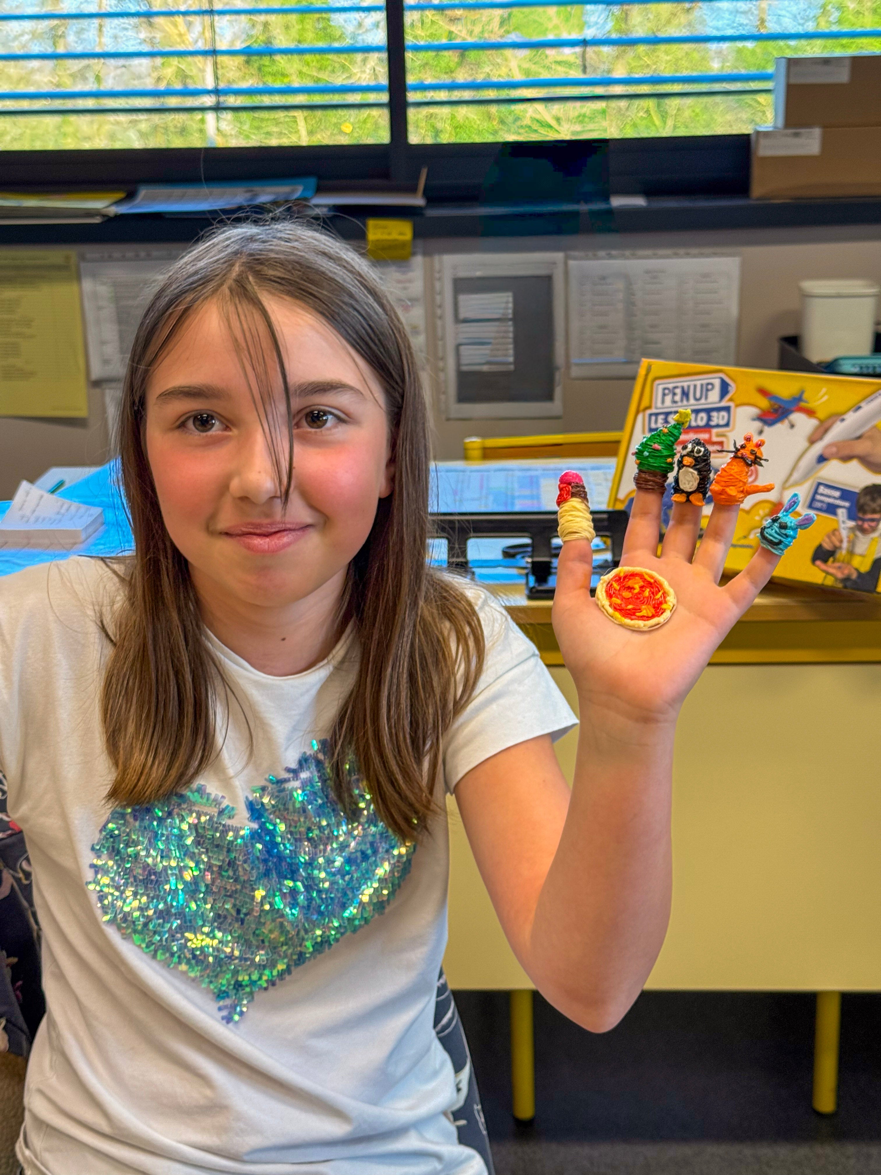 Une fille souriante montre sa main ouverte avec cinq marionnettes de doigt et une mini pizza en fil 3D, la boite Pen'Up visible derriere elle.