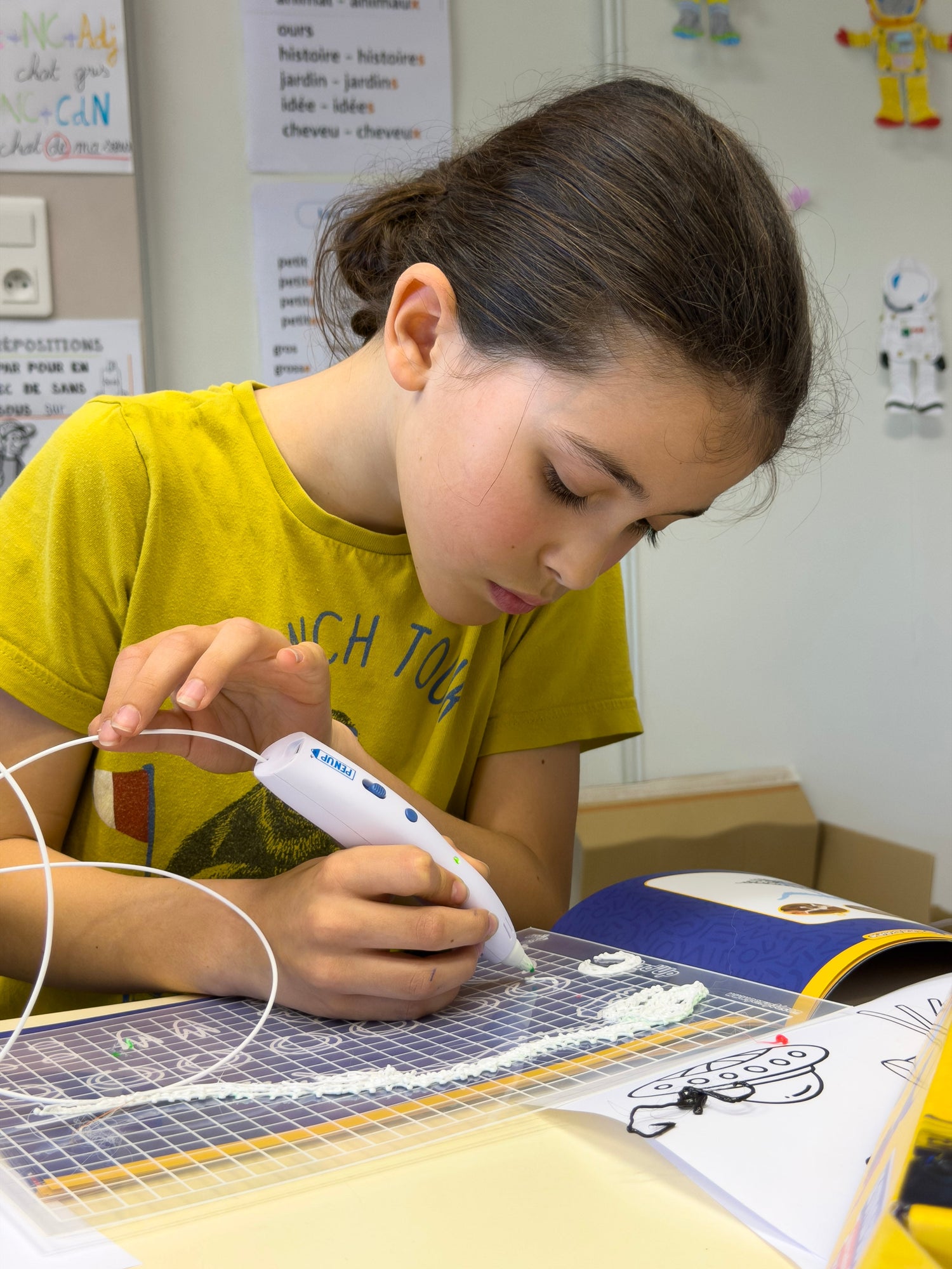 Une fille concentree trace au stylo 3D Pen'Up sur le tapis quadrille transparent pose sur le livret.