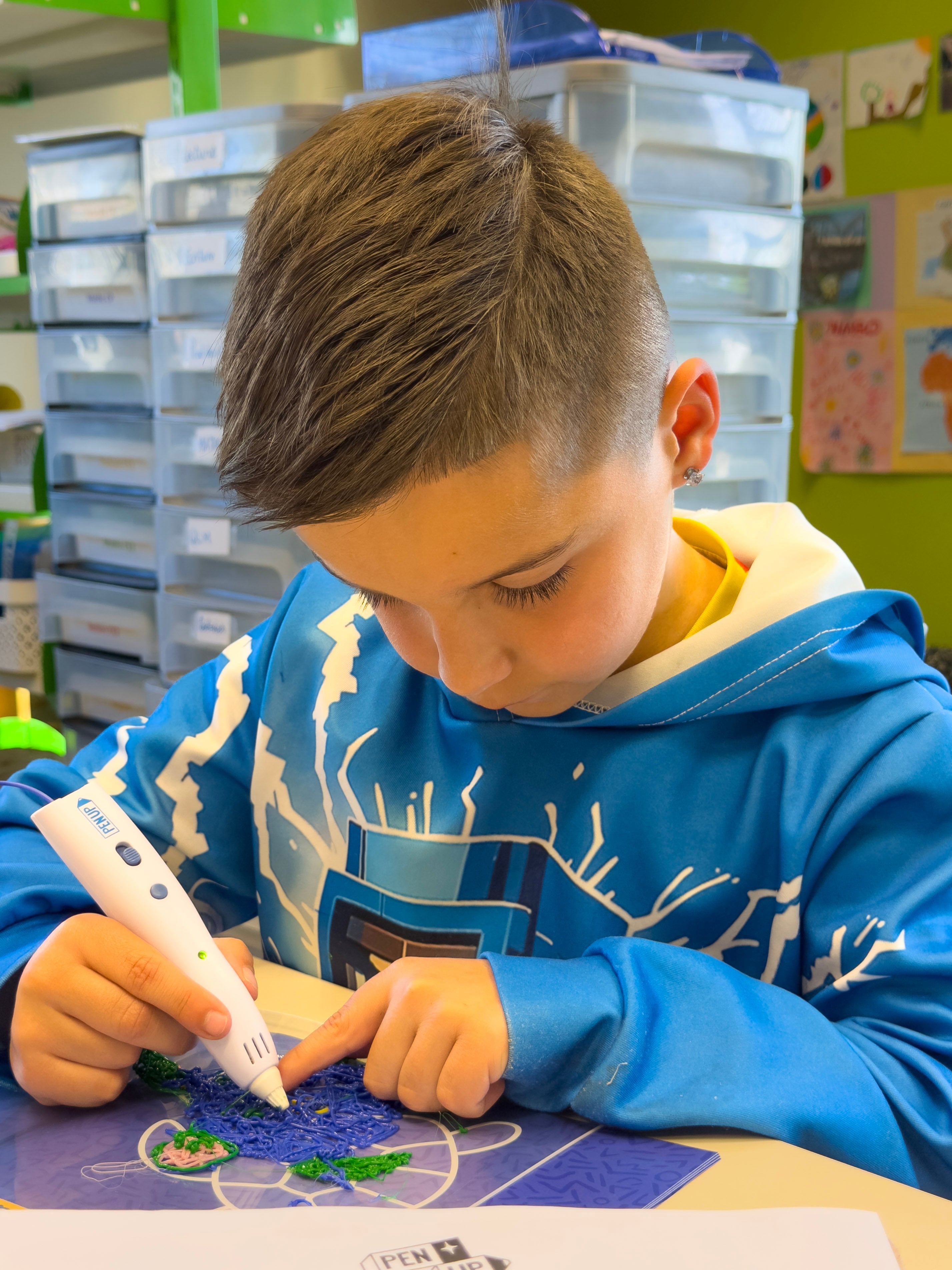Un garcon concentre remplit une tortue en filament bleu et vert avec le stylo 3D Pen'Up sur le tapis de modele.
