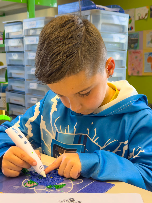 Un garcon concentre remplit une tortue en filament bleu et vert avec le stylo 3D Pen'Up sur le tapis de modele.