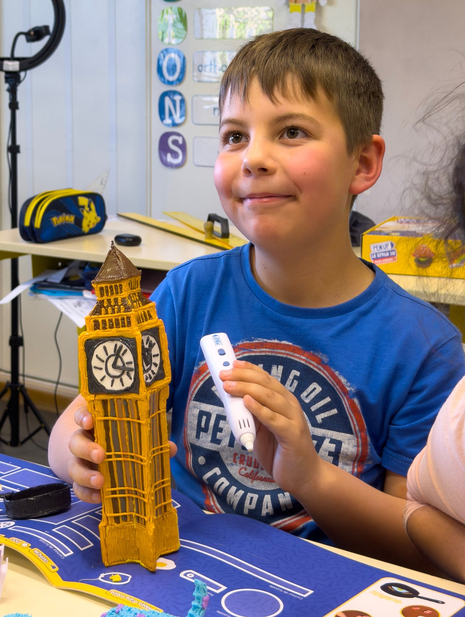 Un garcon souriant tient une grande tour Big Ben jaune realisee au stylo 3D Pen'Up dans une salle de classe.