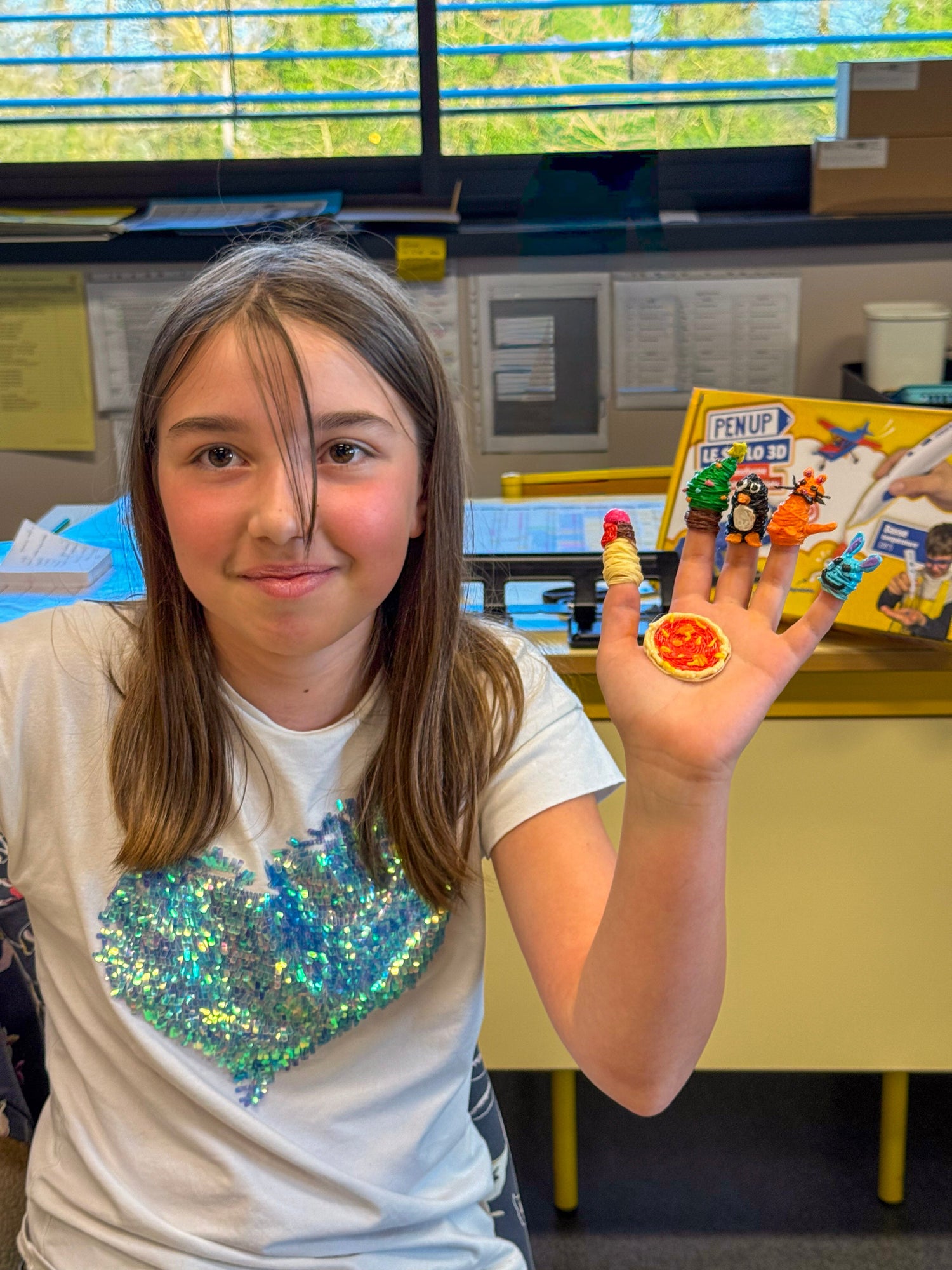 Une fille souriante montre sa main ouverte avec cinq marionnettes de doigt et une mini pizza en fil 3D, la boite Pen'Up visible derriere elle.