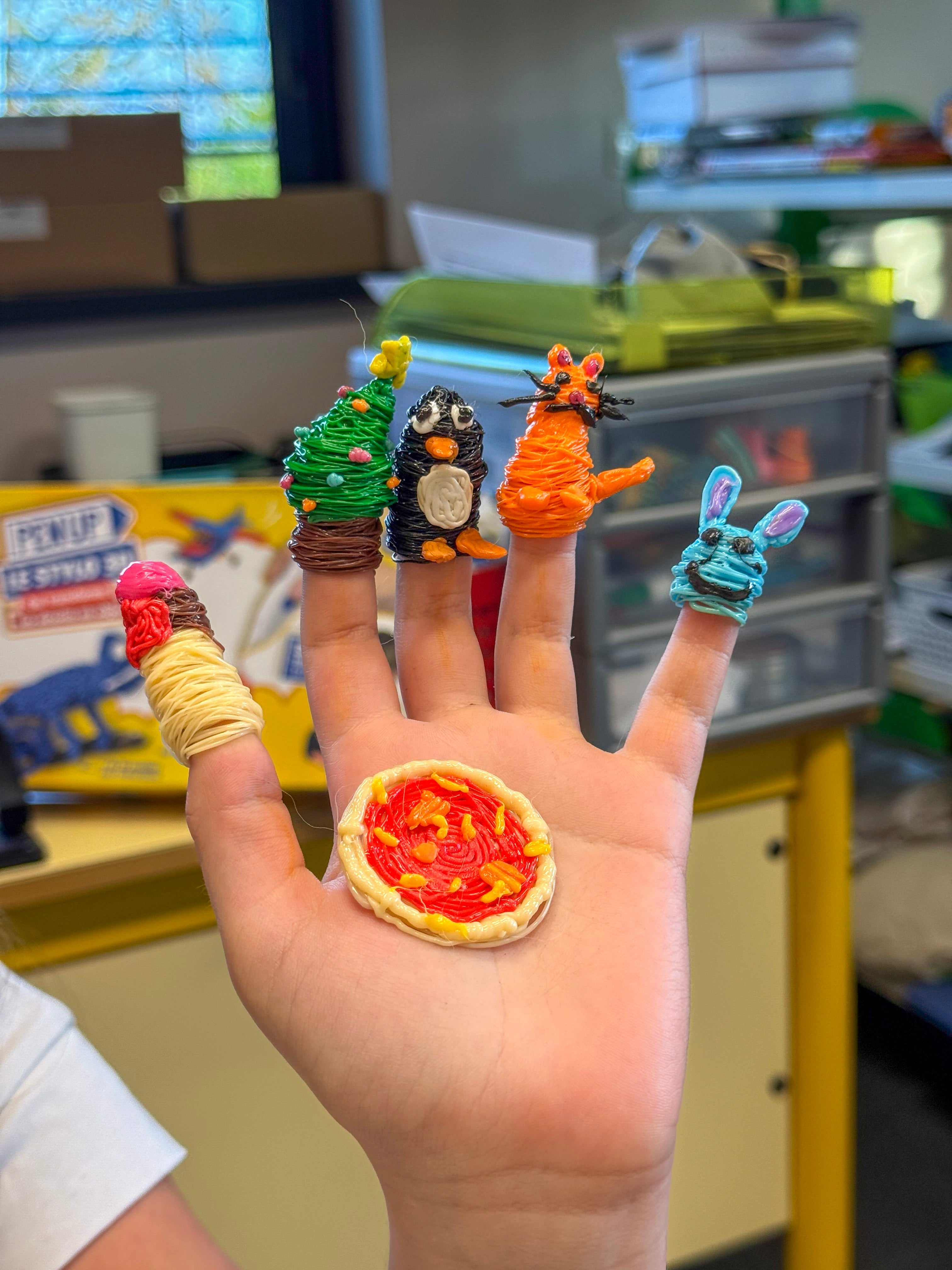 Gros plan sur la main ouverte d'un enfant portant cinq marionnettes de doigt en fil 3D (sapin, pingouin, tigre, lapin, glace) et une mini pizza posée sur la paume.
